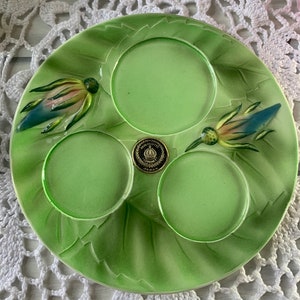 May include: A green ceramic plate with three indented circles and two raised floral designs. The plate has a gold sticker with the words "Hand Painted Japan" on it.
