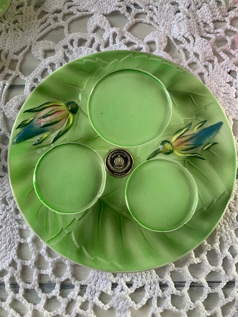 May include: A green ceramic plate with three indented circles and two raised floral designs. The plate has a gold sticker with the words "Hand Painted Japan" on it.
