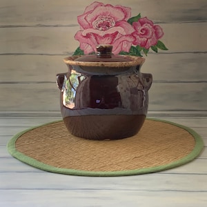 May include: A brown glazed ceramic crock with a lid. The crock has a brown and white glaze and is sitting on a round woven mat with a green border.