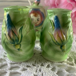 May include: A pair of green ceramic salt and pepper shakers with floral designs. The shakers feature a raised, three-dimensional flower design with blue, pink, and yellow accents.