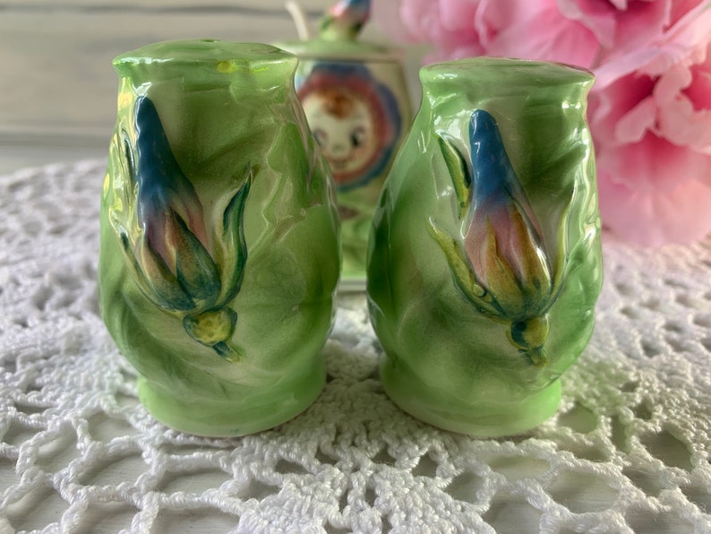 May include: A pair of green ceramic salt and pepper shakers with floral designs. The shakers feature a raised, three-dimensional flower design with blue, pink, and yellow accents.