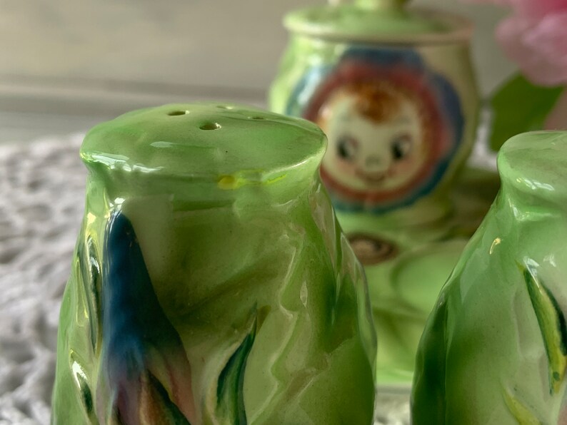May include: A set of three green ceramic salt and pepper shakers with floral designs. The shakers are decorated with blue, pink, and white flowers.