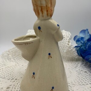 May include: A ceramic figurine of a girl with blonde hair and a white dress with blue polka dots. The girl is holding a small basket in her arm. The figurine is a vintage style and has a rustic look.