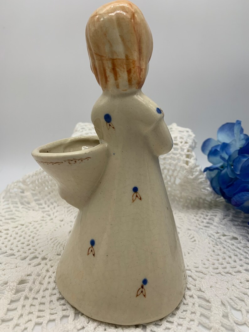 May include: A ceramic figurine of a girl with blonde hair and a white dress with blue polka dots. The girl is holding a small basket in her arm. The figurine is a vintage style and has a rustic look.