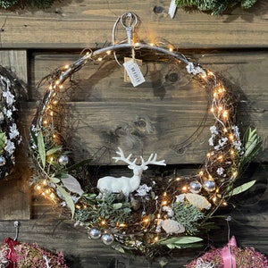 Op de afbeelding: Een decoratieve kerstkrans met een metalen frame, versierd met twinkelende lichtjes, zilveren ornamenten en een wit rendierfiguur. De krans is gemaakt van natuurlijke takken en groen, een feestelijke kerstversiering.