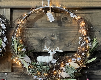May include: A decorative Christmas wreath with a metal frame, adorned with twinkling fairy lights, silver ornaments, and a white reindeer figurine. The wreath is made of natural twigs and greenery, creating a festive holiday decoration.