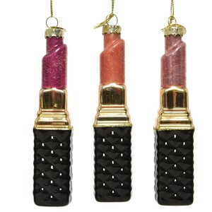 Könnte beinhalten: Drei gläserne Weihnachtsbaumschmuckstücke in Form von Lippenstifttuben. Die Lippenstifttuben sind in den Farben Rosa, Rot und Nude gehalten. Die Tuben haben ein schwarzes und goldenes Design.