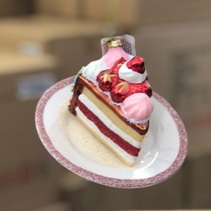 Könnte beinhalten: Ein gläserner Weihnachtsbaumschmuck in Form eines Stückes Erdbeerkuchen mit rosa Zuckerguss und einer roten Erdbeere obenauf. Der Kuchen befindet sich auf einem weißen Teller mit rosa Rand.