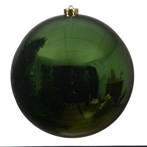 Könnte beinhalten: Eine große, runde, grüne Weihnachtskugel mit glänzender, reflektierender Oberfläche. Die Kugel hat eine goldfarbene Metallkappe zum Aufhängen. Die Oberfläche spiegelt einen Raum mit Stativ, Möbeln und einem Weihnachtsbaum wider.