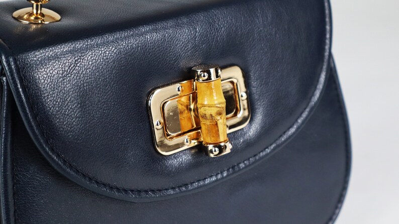 K&ouml;nnte beinhalten: Eine dunkelblaue Ledertasche mit einem goldenen Verschluss und einem Bambus-Knopf.