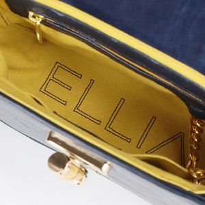 K&ouml;nnte beinhalten: Innenansicht einer blauen Handtasche mit gelbem Futter. Das Futter ist mit dem Wort "ELLIA" in schwarzem Faden gestickt.