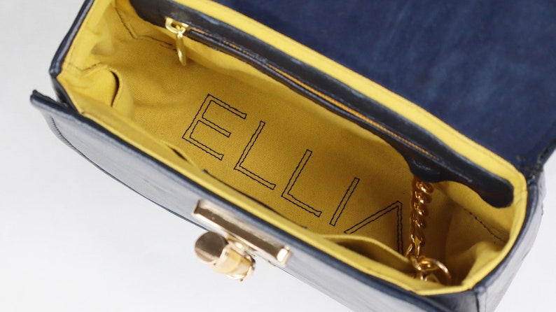 K&ouml;nnte beinhalten: Innenansicht einer blauen Handtasche mit gelbem Futter. Das Futter ist mit dem Wort "ELLIA" in schwarzem Faden gestickt.