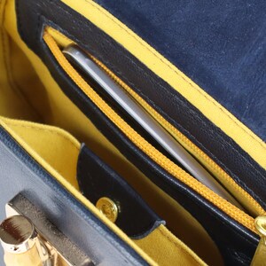 K&ouml;nnte beinhalten: Das Innere einer blauen Ledertasche mit gelber Innenausstattung. Eine Rei&szlig;verschlusstasche mit einem silbernen Telefon darin ist sichtbar.