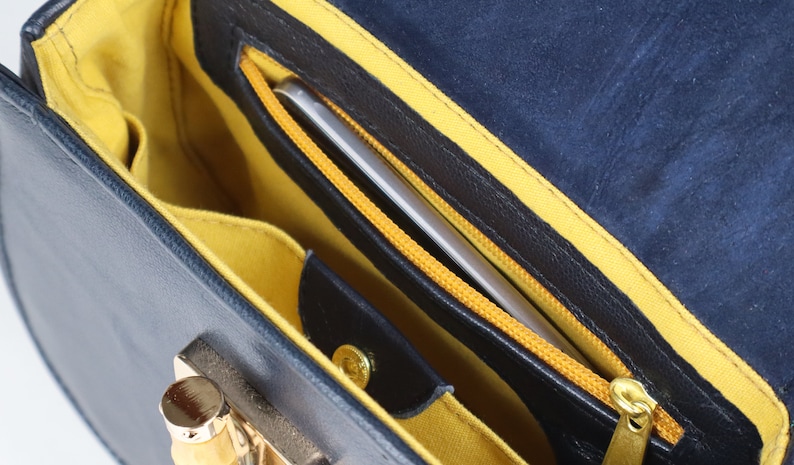 K&ouml;nnte beinhalten: Das Innere einer blauen Ledertasche mit gelber Innenausstattung. Eine Rei&szlig;verschlusstasche mit einem silbernen Telefon darin ist sichtbar.