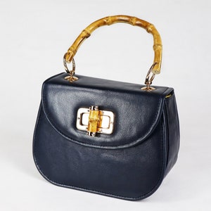 K&ouml;nnte beinhalten: Eine dunkelblaue Ledertasche mit einem Bambusgriff und einem goldenen Verschluss. Der Verschluss hat ein kleines St&uuml;ck Bambus in der Mitte.