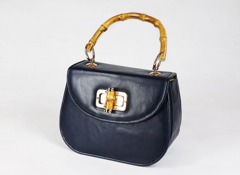 K&ouml;nnte beinhalten: Eine dunkelblaue Ledertasche mit einem Bambusgriff und einem goldenen Verschluss. Der Verschluss hat ein kleines St&uuml;ck Bambus in der Mitte.