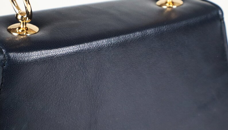 K&ouml;nnte beinhalten: Eine dunkelblaue Ledertasche mit goldfarbenem Henkel und Beschl&auml;gen.