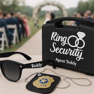 以下が含まれることがあります： テディという名前がアームに書かれた黒いサングラス、リングセキュリティの文字が入った黒いケース、チェーン付きのバッジ。ケースにはダイヤモンドの指輪のグラフィックがあります。アイテムは、結婚式のセレモニーを背景にテーブルに置かれています。