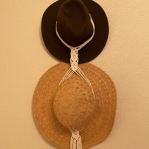 May include: Two hats hanging on a macrame hat rack. The top hat is brown felt and the bottom hat is a woven straw hat.