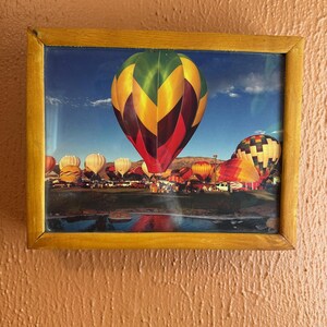 May include: A framed print featuring a vibrant display of hot air balloons. The central balloon is a mix of green, yellow, and red, with others in various colors. The scene includes a body of water and a blue sky.