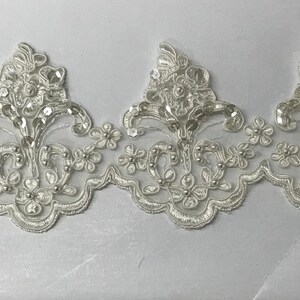 May include: White lace trim with a delicate floral pattern and small sequins. The trim has a scalloped edge and is perfect for adding a touch of elegance to any project.