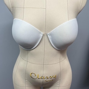 Puede incluir: Un sujetador blanco sin tirantes expuesto en un maniquí beige. El sujetador tiene un escote curvo y está hecho de una tela lisa. El maniquí tiene líneas de cuadrícula y el texto dorado "Classy forms".