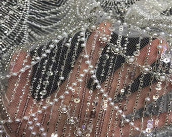May include: Close-up of a white beaded and sequined fabric. The fabric has a delicate, sheer texture with rows of small white pearls and silver sequins.