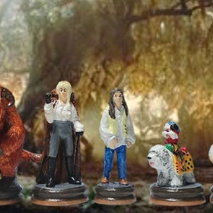 Op de afbeelding: Een set van zes handgeschilderde beeldjes, waaronder een bruin, harig wezen met hoorns, een figuur in een wit shirt en een bruine jas, een figuur in een gele vest en blauwe jeans, een witte hond met een kleurrijk figuur op zijn rug, en twee figuren in bruine shirts. De beeldjes zijn geplaatst tegen een wazige achtergrond van een bos.