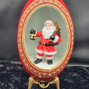 May include: A red, jeweled egg with a gold stand, featuring a Santa Claus figurine inside. The Santa Claus figurine is holding a lantern and a bag of gifts.