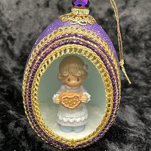 May include: Purple glitter egg ornament with gold trim and a small figurine of a girl holding a heart-shaped cookie inside.