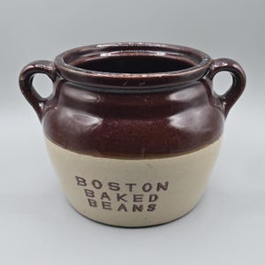 May include: A two-handled ceramic bean pot with a glossy dark brown upper section and a matte beige lower section. The words BOSTON BAKED BEANS are embossed in dark brown lettering on the front of the pot, which features a wide opening.
