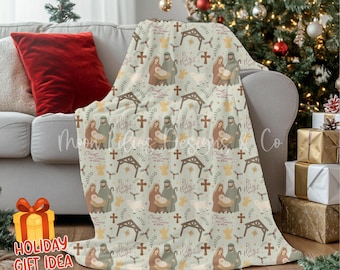 Puede incluir: Una manta verde claro con un patrón repetido de una escena de la natividad, cruces y estrellas. La manta está sobre un sofá gris con un cojín rojo. Un árbol de Navidad está en el fondo. Las palabras "Holiday Gift Idea" están en la esquina inferior izquierda.