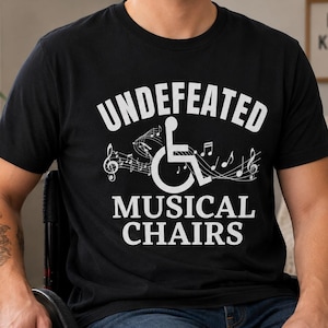 May include: Black t-shirt with the words "Undefeated Musical Chairs" in white, along with a wheelchair symbol and musical notes. The shirt is worn by a person in a wheelchair.