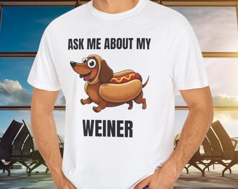 May include: White t-shirt with the text "ASK ME ABOUT MY WEINER." The shirt features a cartoon illustration of a dachshund dog inside a hot dog bun, with a smiling face and a hot dog inside.