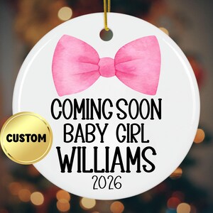 May include: A white, round Christmas ornament with a pink watercolor bow design. The ornament has the text "COMING SOON BABY GIRL WILLIAMS 2026". A gold "CUSTOM" sticker is on the left side.