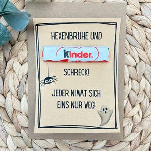 Peut inclure: Une carte rectangulaire en papier kraft avec une barre de chocolat Kinder fixée au centre. La carte présente une bordure noire dessinée à la main et le texte : HEXENBRÜHE UND SCHRECK! JEDER NIMMT SICH EINS NUR WEG! Elle inclut une illustration de fantôme et d'araignée.