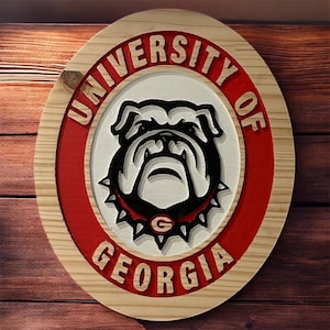May include: A wooden, layered sign featuring the University of Georgia logo. The sign is circular with a red and white border, and the words "UNIVERSITY OF GEORGIA" are carved into the wood. The central image is a bulldog.