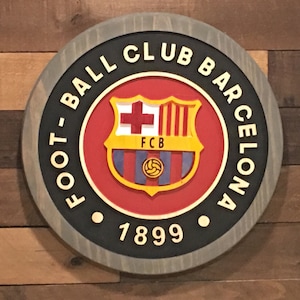 May include: A round wooden sign with a black border and the text "FOOTBALL CLUB BARCELONA 1899". The center of the sign features the FC Barcelona crest, which is a red shield with a yellow border and a blue and red striped pattern. The crest also includes a yellow cross, a yellow ball, and the text "FCB".