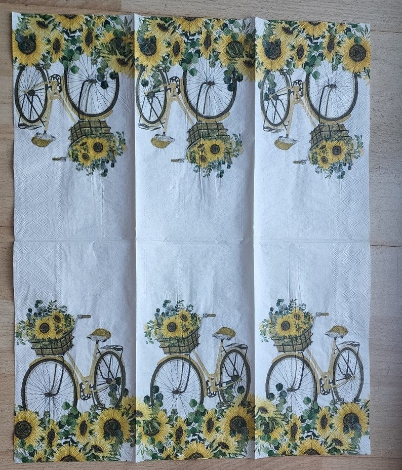 Puede incluir: Una servilleta de papel blanca con un patr&oacute;n repetido de girasoles amarillos y una bicicleta con una cesta llena de girasoles.