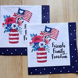 May include: Two white paper napkins with a blue and white polka dot border. The napkins feature a red, white and blue American flag, a mason jar filled with red, white and blue flowers, and the text "Friends Family Freedom".