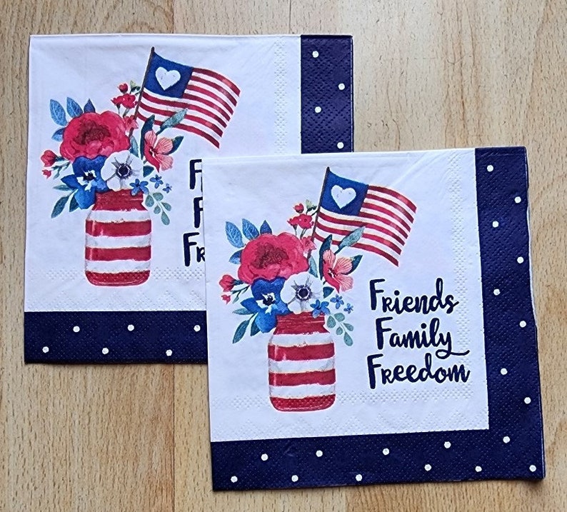 May include: Two white paper napkins with a blue and white polka dot border. The napkins feature a red, white and blue American flag, a mason jar filled with red, white and blue flowers, and the text "Friends Family Freedom".
