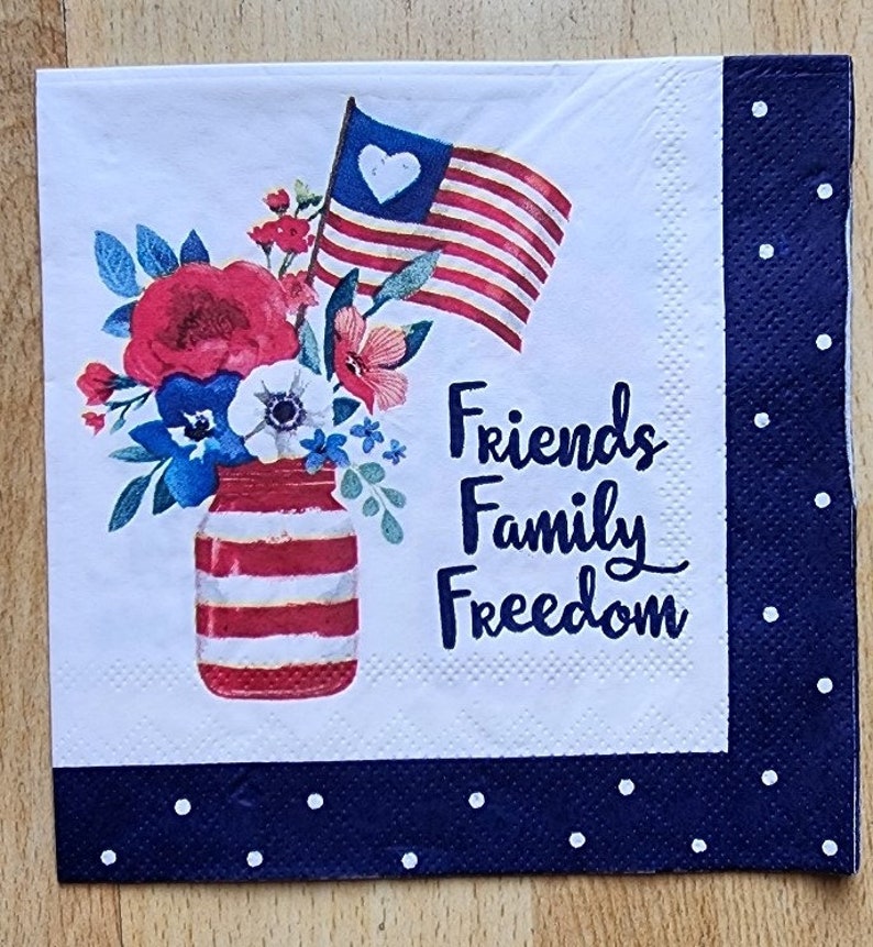 May include: A white paper napkin with a red, white and blue striped mason jar filled with red, white and blue flowers. The jar has a red, white and blue striped lid. An American flag with a white heart in the top left corner is behind the flowers. The text "Friends Family Freedom" is printed on the napkin.