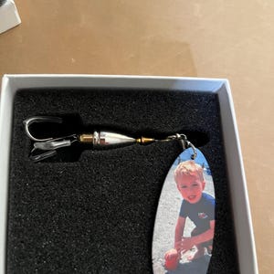 Könnte beinhalten: Ein silberfarbener Angelköder mit einem Foto eines kleinen Kindes auf einem tropfenförmigen Anhänger. Der Köder befindet sich in einer weißen Box mit schwarzer Schaumstoffpolsterung.