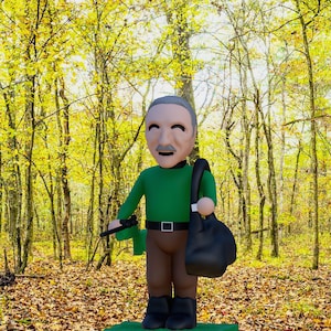 May include: A figurine of an older person with gray hair and a mustache, wearing a green long-sleeved shirt, a black belt, brown pants, and black boots. The figure holds a black bag over one shoulder and a small black object in the other hand.