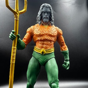 Puede incluir: Una figura de acción detallada de Aquaman, un superhéroe de DC Comics. La figura lleva un traje verde y naranja con un cinturón dorado y sostiene un tridente dorado. La figura tiene una barba larga y oscura y el pelo largo y oscuro.