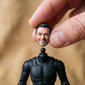 Può includere: Una testa di action figure dettagliata con un volto maschile realistico, capelli scuri, barba leggera ed espressione sorridente, posizionata sopra un busto in plastica nera con un connettore a sfera per il collo.