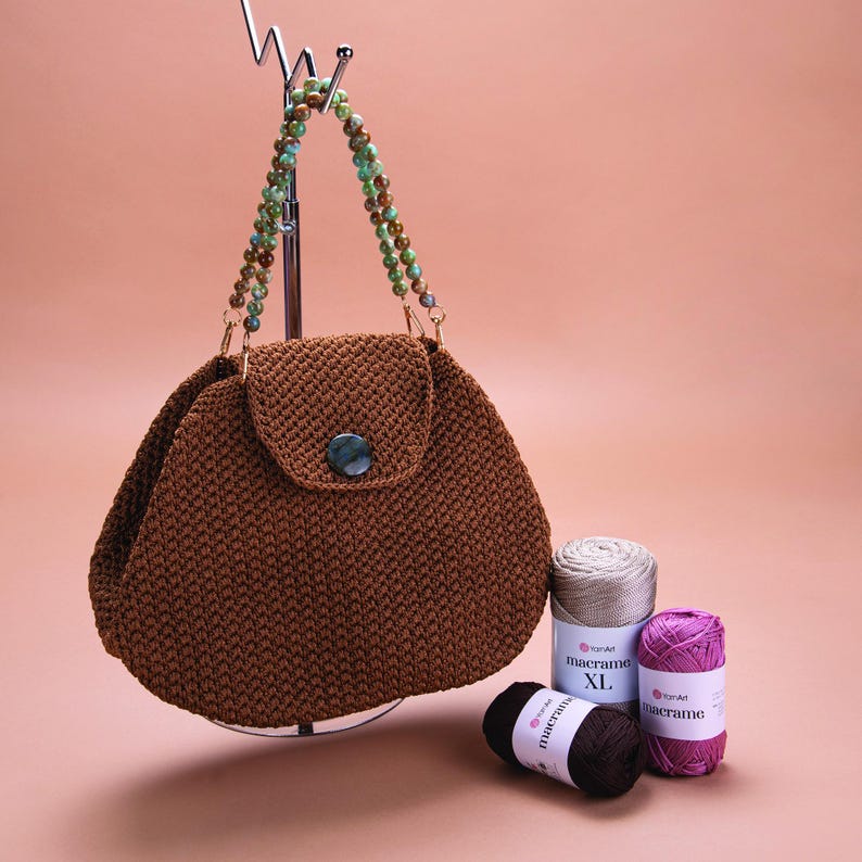 May include: A brown crocheted handbag with a flap closure and a beaded strap. The bag is hanging from a metal stand. Three balls of yarn are in the foreground, labeled "macrame XL", "macrame", and "macrame".