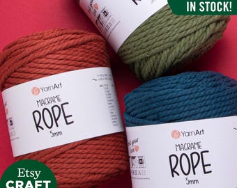 May include: Three spools of 5mm macrame rope in rust, forest green, and teal blue. The spools are labeled "YarnArt Macrame Rope 5mm". The text "All Colors In Stock!" is displayed in a green box.