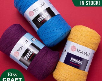 May include: Four spools of YarnArt Ribbon yarn in blue, purple, yellow, and orange. The spools are on a red background. The text "ALL COLORS IN STOCK!" is in green text on a white background.