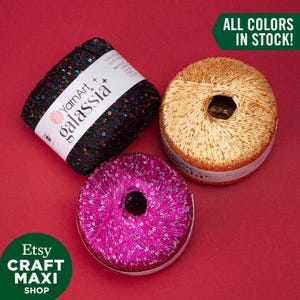 May include: Three spools of YarnArt Galassia yarn in black, gold, and pink, each with sequins. The black yarn is angled, while the gold and pink spools are circular. The image includes the text "ALL COLORS IN STOCK!" and "Etsy CRAFT MAXI SHOP."